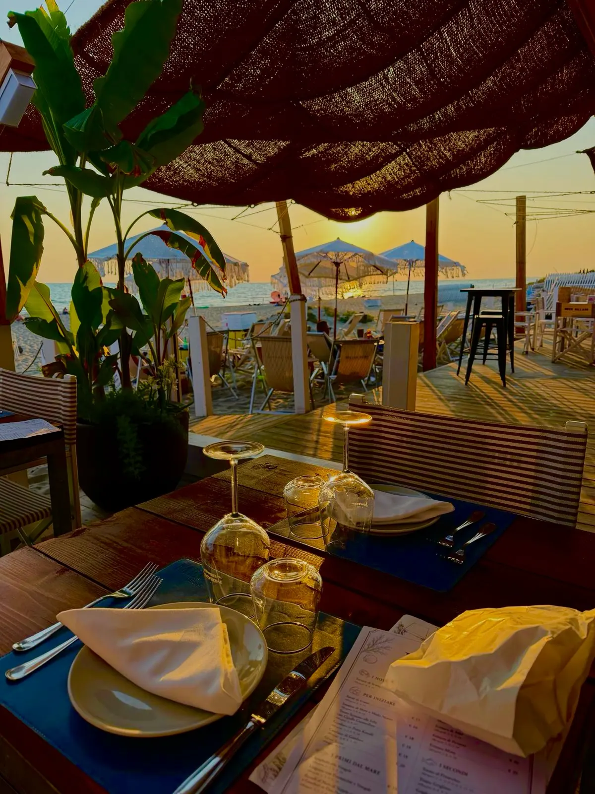 Il sole al tramonto sul mare visto da sotto le tende, tra gli ombrelloni di paglia